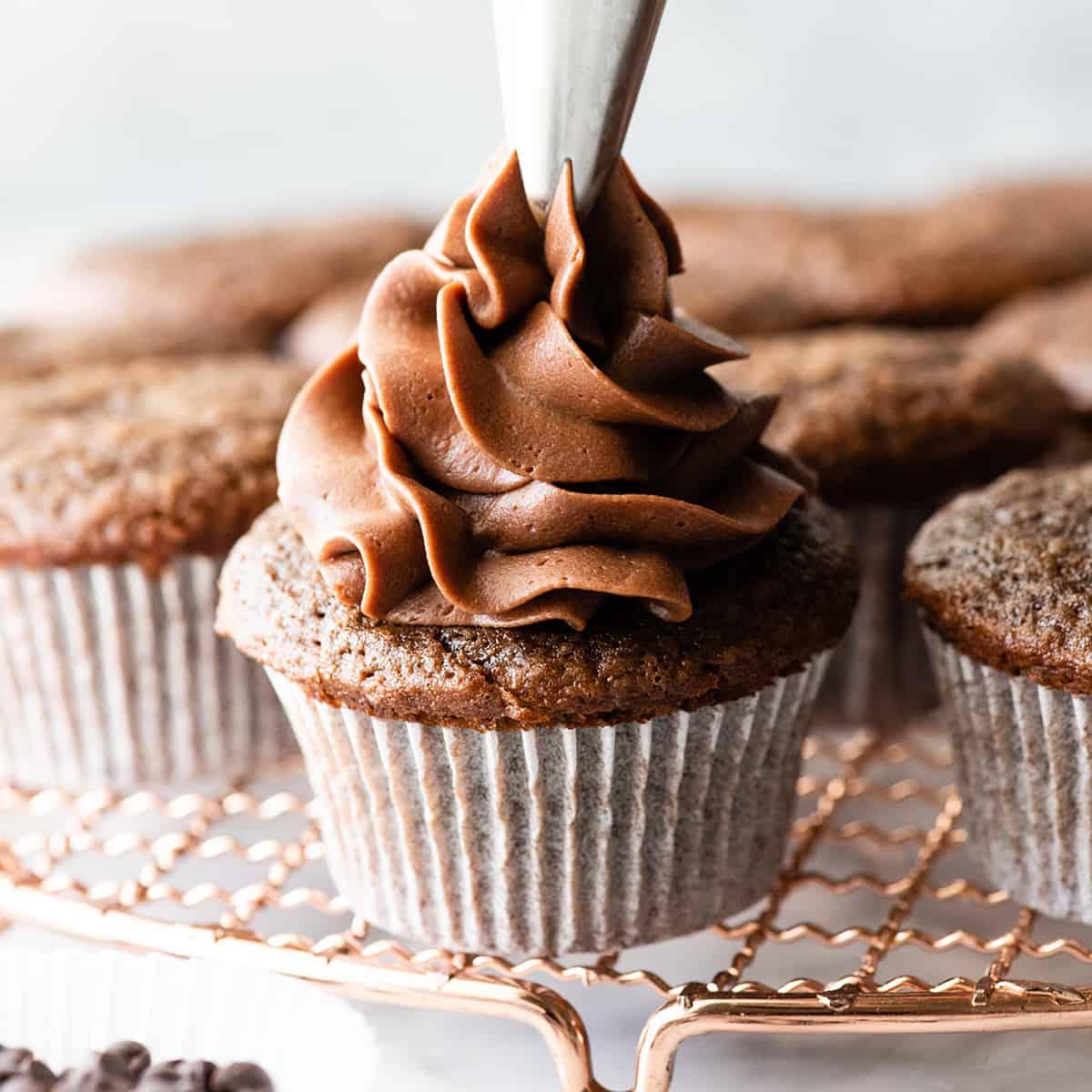 Αυτή η συνταγή για σοκολατένια cupcakes είναι εύκολη στην παρασκευή από το μηδέν με απλά υλικά. Είναι τα καλύτερα, ζουμερά σοκολατένια cupcakes με πλούσια γεύση σοκολάτας και τέλεια υφή. Μπροστινή φωτογραφία που δείχνει γλάσο να πιέζεται πάνω σε ένα σοκολατένιο cupcake