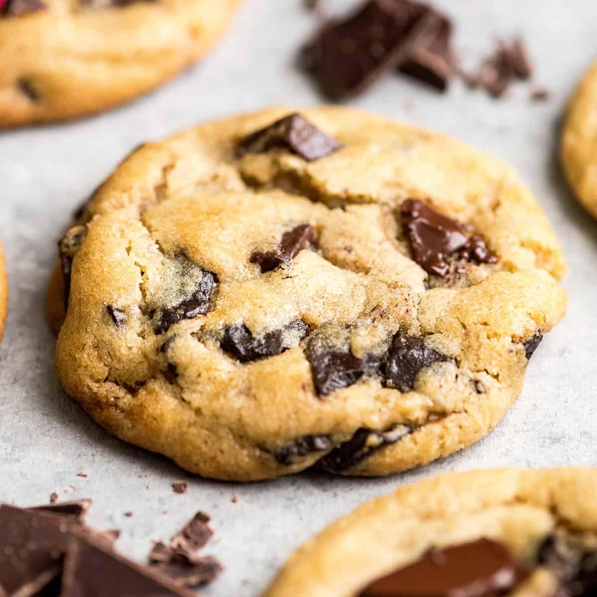 Αυτή είναι η καλύτερη συνταγή για chocolate chip cookies που έχετε δοκιμάσει ποτέ. Χωρίς περίεργα υλικά, χωρίς χρόνο ψύξης, κ.λπ. Απλά ένα απλό, εύκολο στην παρασκευή του, απίστευτα νόστιμο, ζυμαρένιο αλλά πλήρως ψημένο, cookie με κομματάκια σοκολάτας που βγαίνει τέλειο κάθε φορά! #cookies #chocolatechipcookies #baking #recipe #chocolate Προβολή από το πλάι της καλύτερης συνταγής chocolate chip cookie ποτέ, ένα cookie με λιωμένα κομματάκια σοκολάτας περιτριγυρισμένο από άλλα τρία cookies και ψιλοκομμένη σοκολάτα