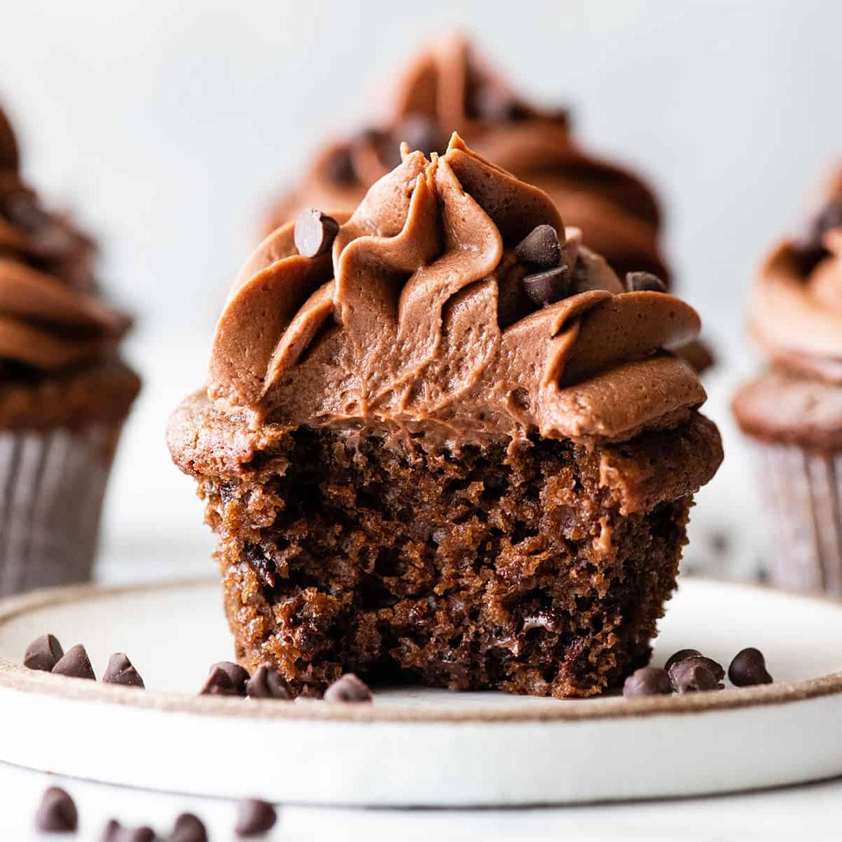 Αυτή η συνταγή για σοκολατένια cupcakes είναι εύκολη στην παρασκευή από το μηδέν με απλά υλικά. Είναι τα καλύτερα, ζουμερά σοκολατένια cupcakes με πλούσια γεύση σοκολάτας και τέλεια υφή. Μπροστινή φωτογραφία ενός σοκολατένιου cupcake με μια δαγκωνιά, σε ένα πιάτο