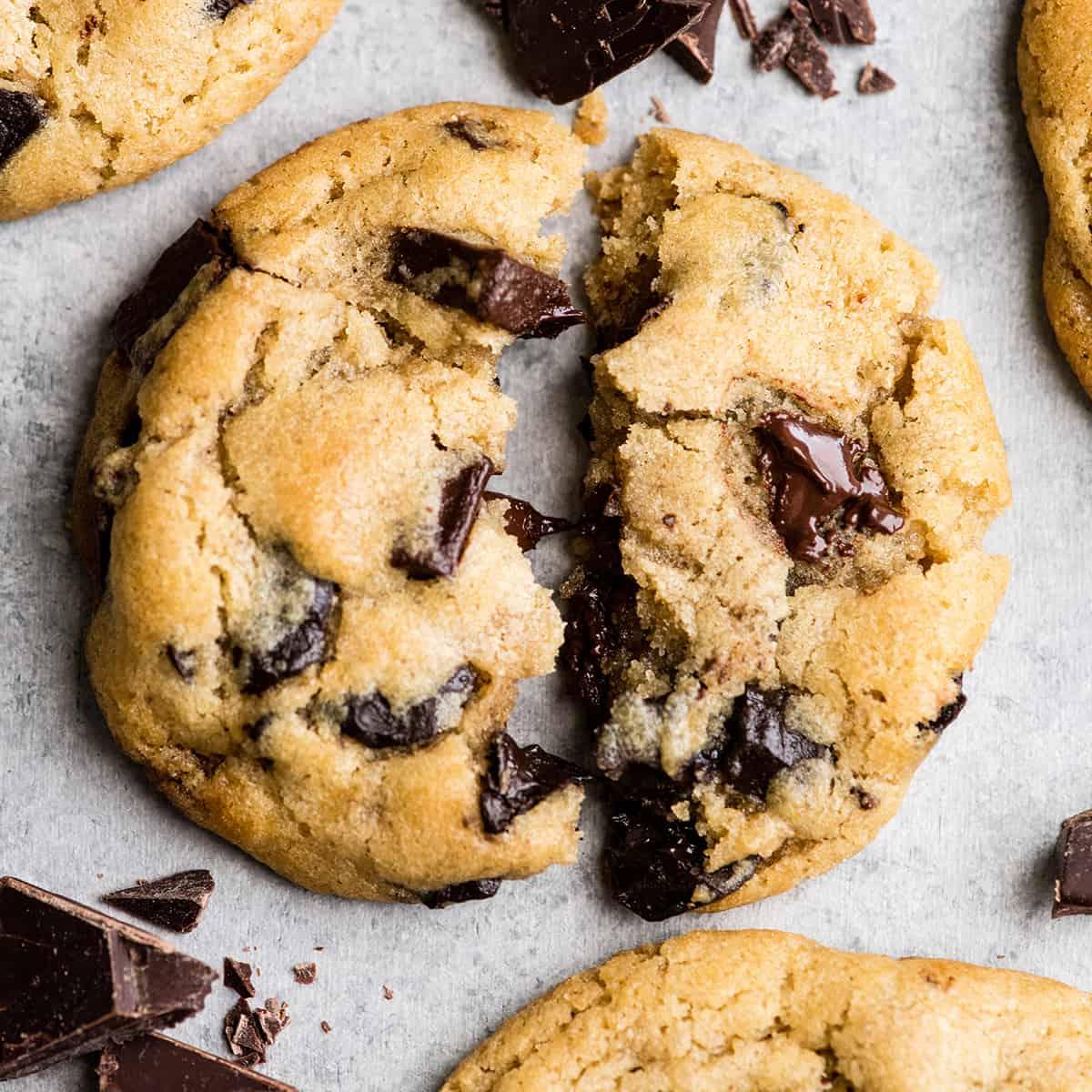 Αυτή είναι η καλύτερη συνταγή για chocolate chip cookies που έχετε δοκιμάσει ποτέ. Χωρίς περίεργα υλικά, χωρίς χρόνο ψύξης, κ.λπ. Απλά ένα απλό, εύκολο στην παρασκευή του, απίστευτα νόστιμο, ζυμαρένιο αλλά πλήρως ψημένο, cookie με κομματάκια σοκολάτας που βγαίνει τέλειο κάθε φορά! #cookies #chocolatechipcookies #baking #recipe #chocolate Προβολή από πάνω ενός chocolate chip cookie κομμένου στη μέση με λιωμένα κομματάκια σοκολάτας, περιτριγυριμένο από άλλα τρία cookies και ψιλοκομμένη σοκολάτα