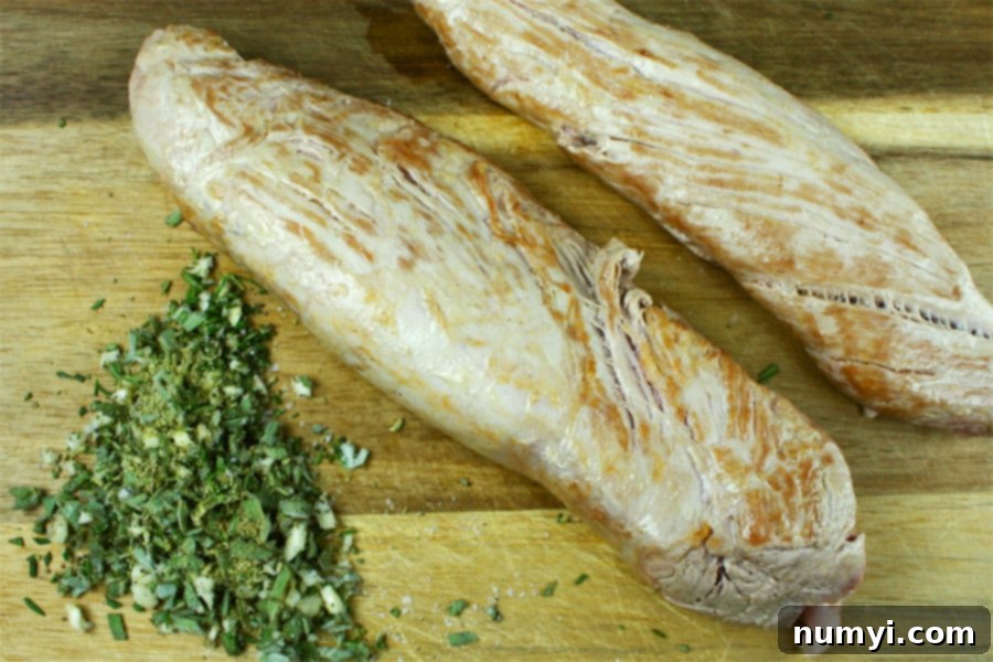 Seared pork tenderloin next to a bowl of freshly chopped Tuscan herbs on a wooden cutting board.