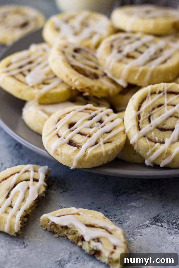 A broken cinnamon roll cookie on a plate next to a stack of whole cookies, showing the delightful cinnamon swirl inside.