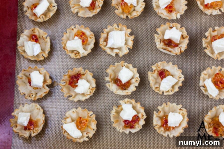 Unbaked phyllo pastry shells arranged on a baking sheet, each filled with a dollop of red pepper jelly and a cube of brie cheese, ready for the oven.