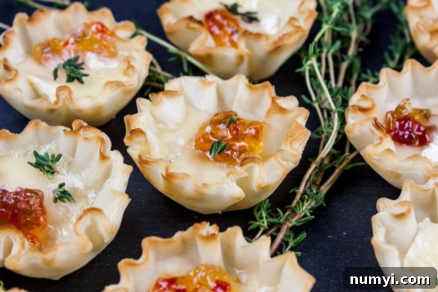A close-up of a single Pepper Jelly Brie Bite, showcasing the melted brie and vibrant red jelly within the crisp phyllo cup, garnished with a tiny thyme leaf.