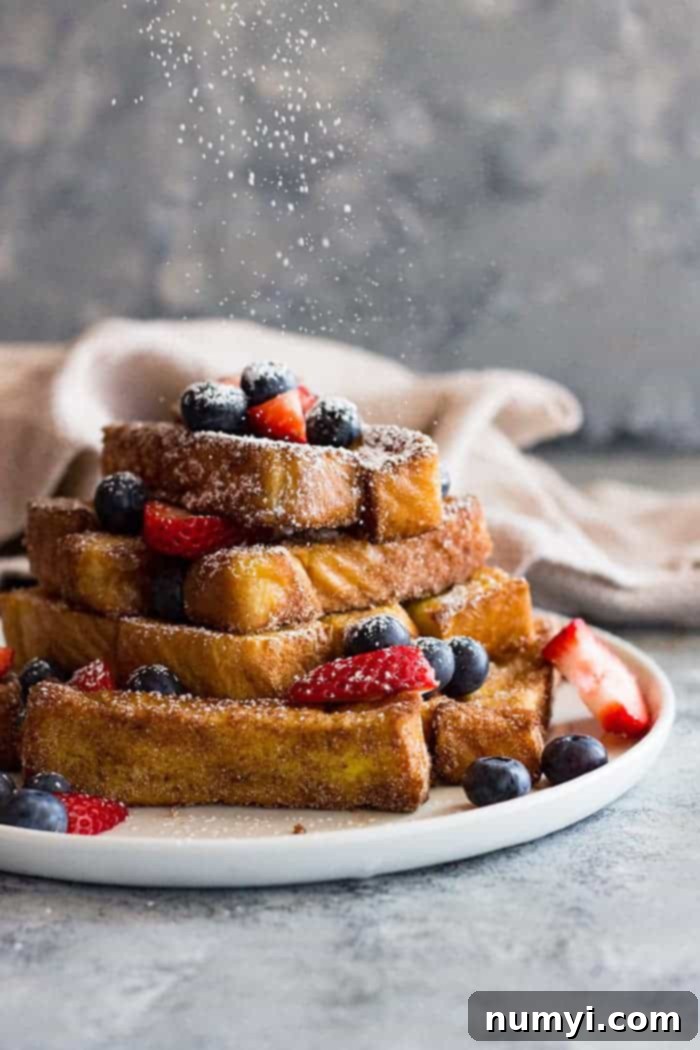A plate piled high with french toast sticks with berries on top. Powdered sugar sprinkling over the top. 