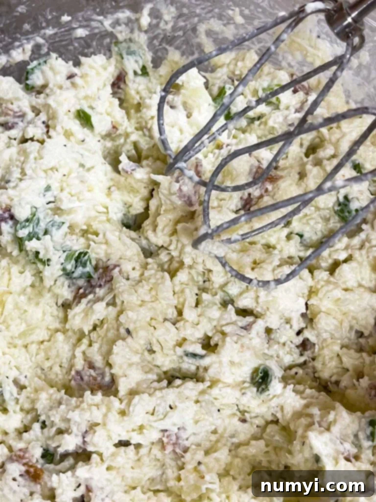 The loaded cauliflower mash in a glass bowl being mixed with an electric handheld mixer for a creamier texture.