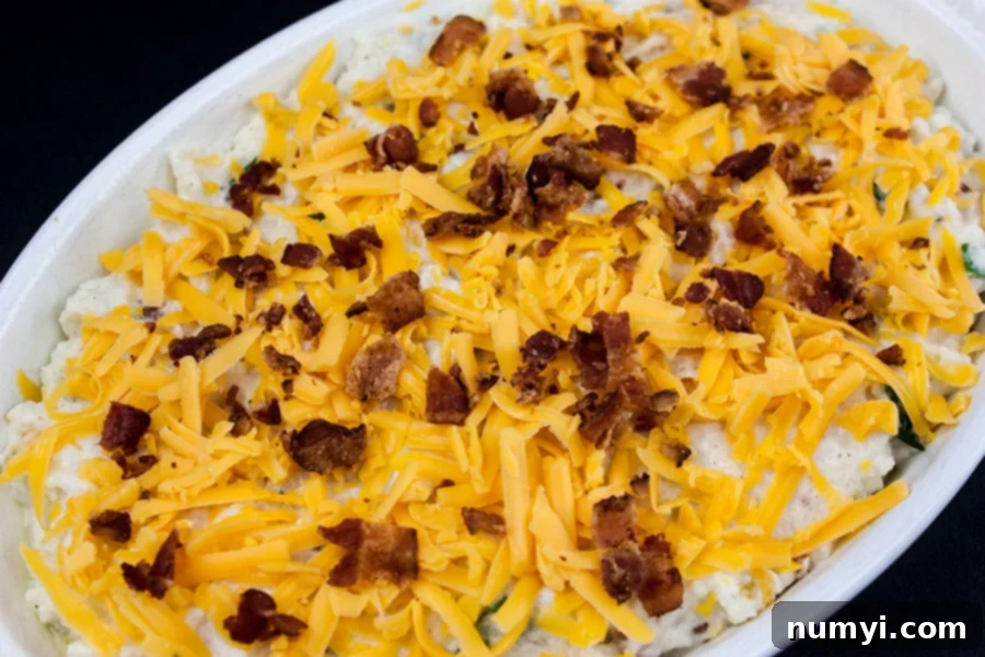 Loaded Cauliflower Mash Casserole in a white casserole dish, topped with shredded cheese and bacon bits, ready for baking.