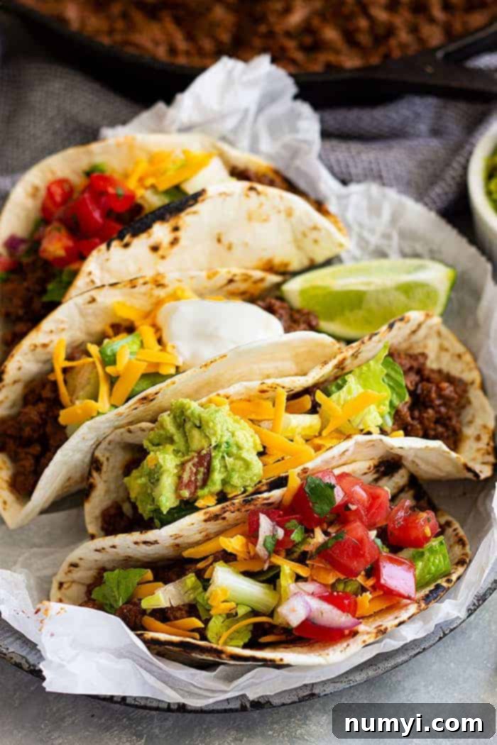 A plate full of ground beef tacos garnished with fresh tomato, creamy guacamole, and shredded cheddar cheese.