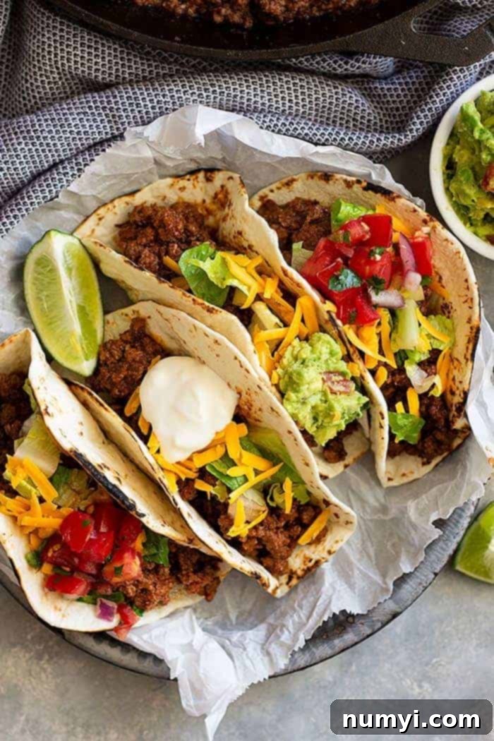 A close-up of a plate of ground beef tacos, garnished with fresh toppings like tomato, creamy guacamole, and shredded cheddar cheese.