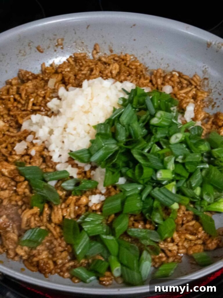Freshly chopped green onions and crunchy water chestnuts being expertly stirred into the seasoned ground chicken mixture in a skillet, adding final touches of flavor and texture.