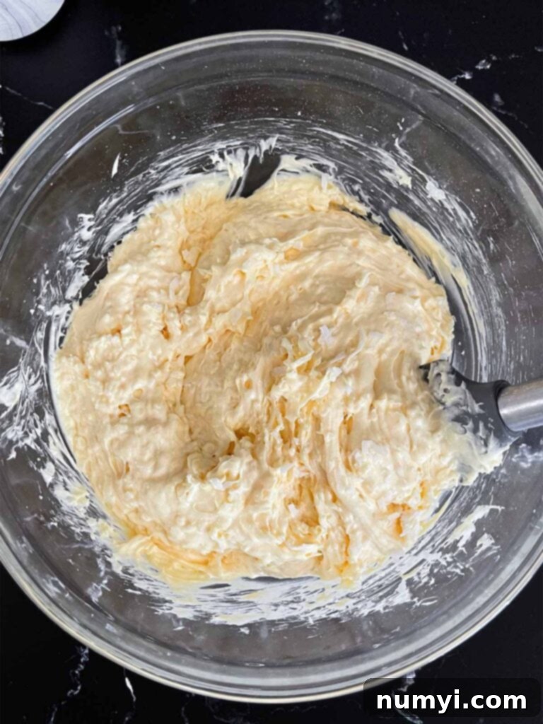 Coconut sheet cake batter in a clear glass mixing bowl on a dark surface, showing flecks of shredded coconut.