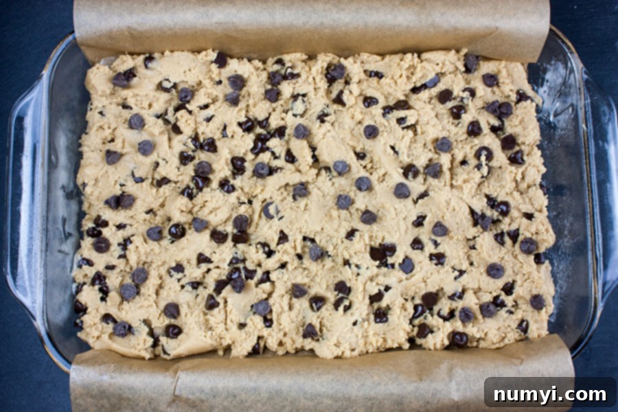 Unbaked chocolate chip cookie dough evenly pressed into a parchment-lined 9x13 baking pan, ready for the oven.