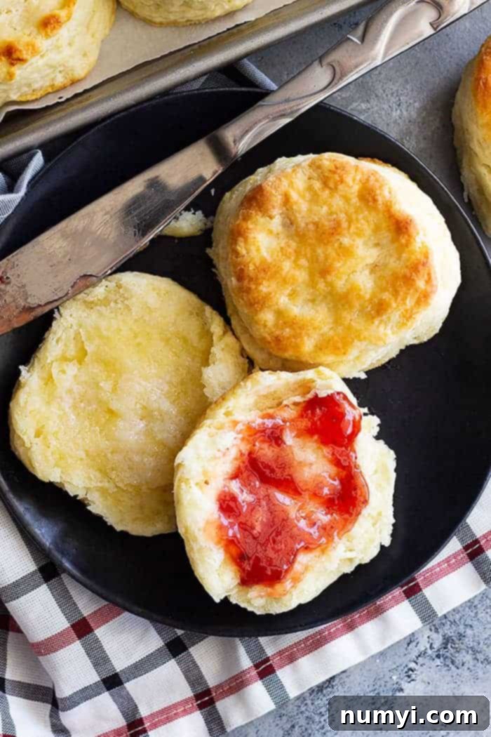 A freshly baked flaky buttermilk biscuit, split open and spread with vibrant strawberry jam, resting on a black plate beside a striped kitchen towel.