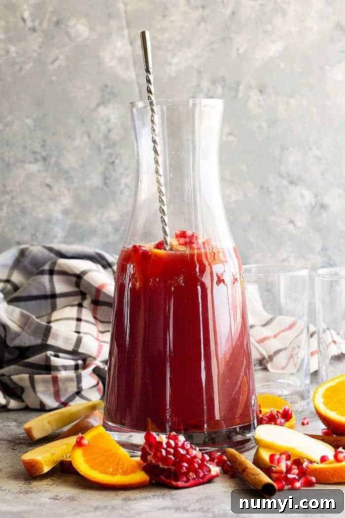 A large serving pitcher of Apple Cider Sangria, brimming with fresh orange and apple slices, and vibrant pomegranate seeds, all surrounded by more seasonal fruit at its base. Perfect for fall and holiday gatherings.