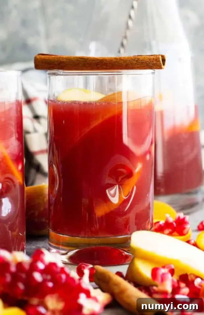 Side view closeup of a tall glass overflowing with Apple Cider Sangria, showcasing vibrant fruit slices and more fresh fruit elegantly arranged on the kitchen counter beside it. The rich, inviting color of the sangria is prominent.