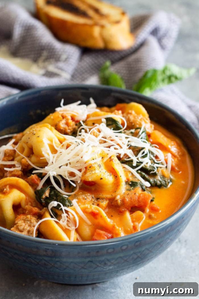 A bowl of creamy sausage and tortellini soup in a blue bowl with a spoon, topped with grated cheese.
