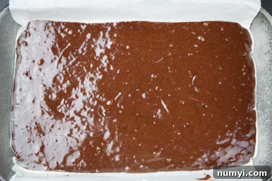 Decadent Turtle Brownies 3 First layer of rich uncooked brownie batter evenly spread in a parchment-lined pan with handles, ready for initial baking.