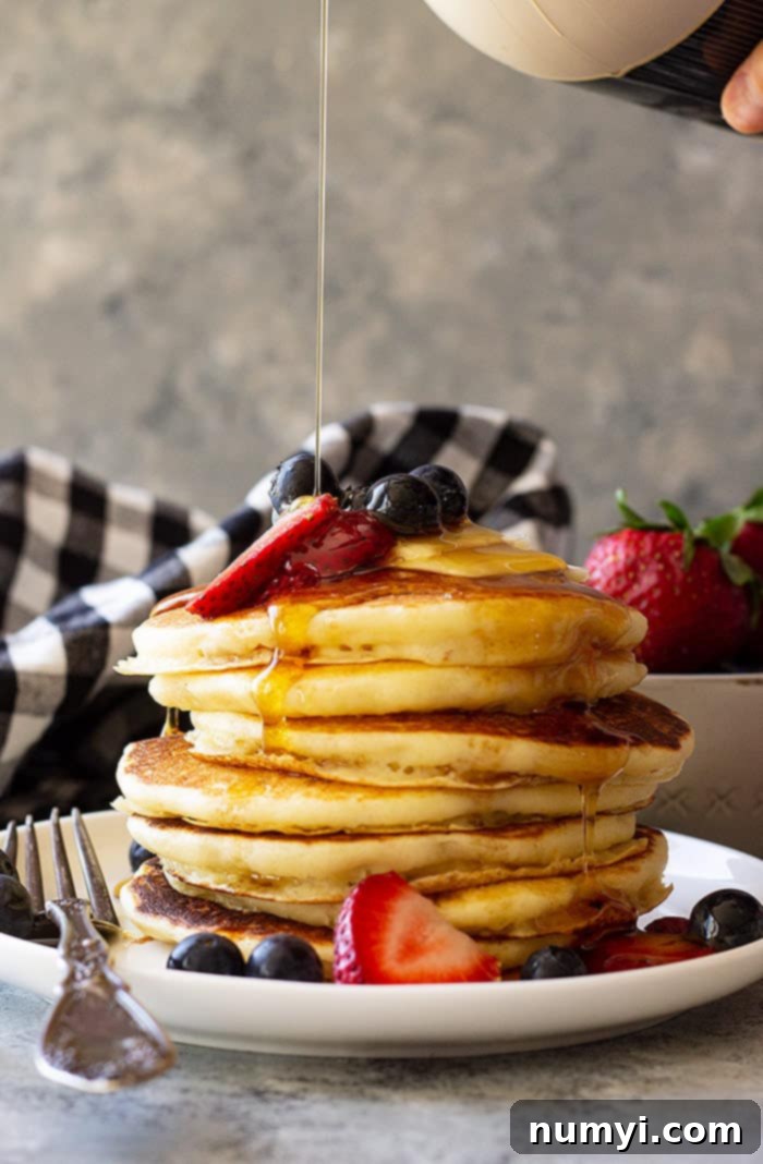 Tall stack of Fluffy Buttermilk Pancakes topped with fresh berries and being drizzled with syrup. 
