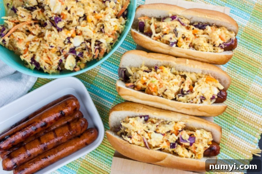 Hot Dogs topped with vibrant Mustard Slaw on a rustic wooden hot dog board, with a bowl of fresh mustard slaw and a platter of perfectly grilled hot dogs in the background.
