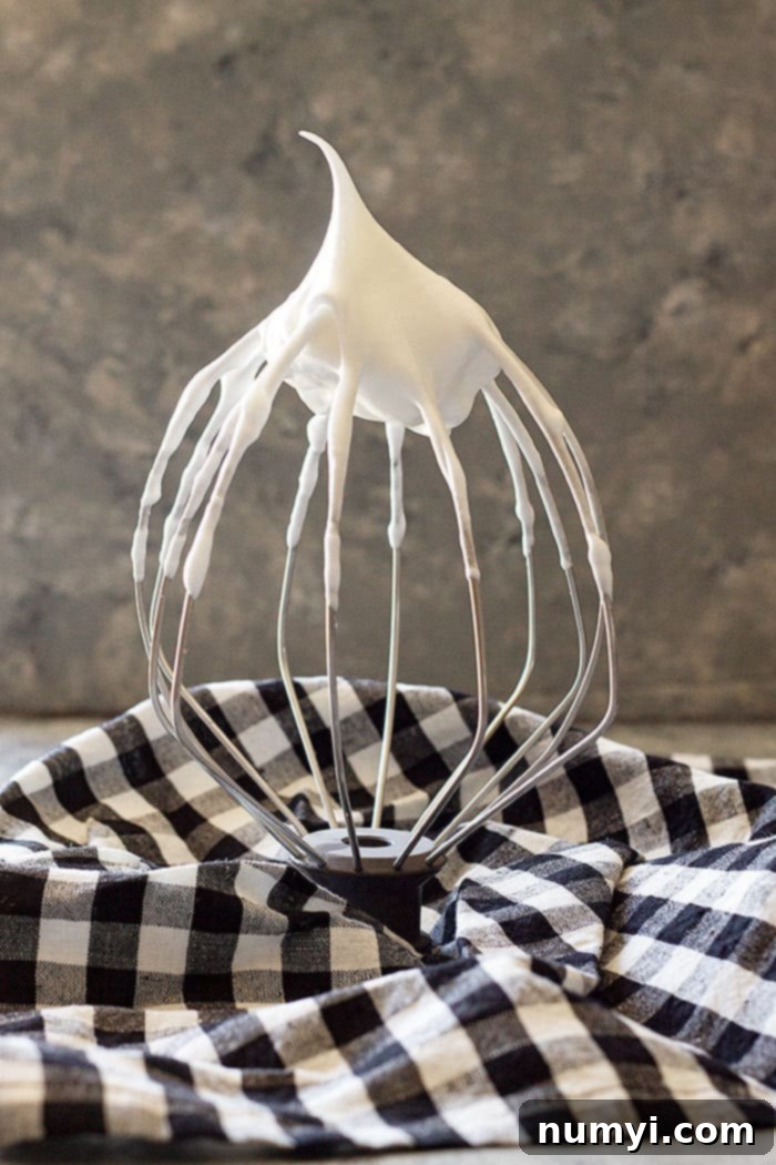 A close-up of an electric mixer whisk attachment covered in beautifully whipped stiff meringue peaks, demonstrating the perfect consistency needed for a successful chocolate meringue pie.