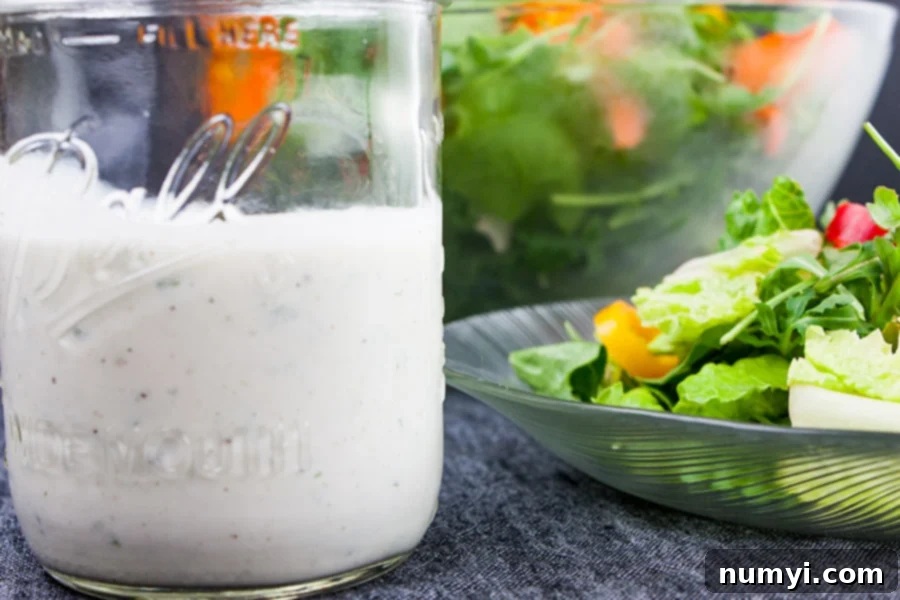 A mason jar filled with creamy Southern buttermilk dressing, with a fresh green salad visible in the softly blurred background, suggesting its perfect use.