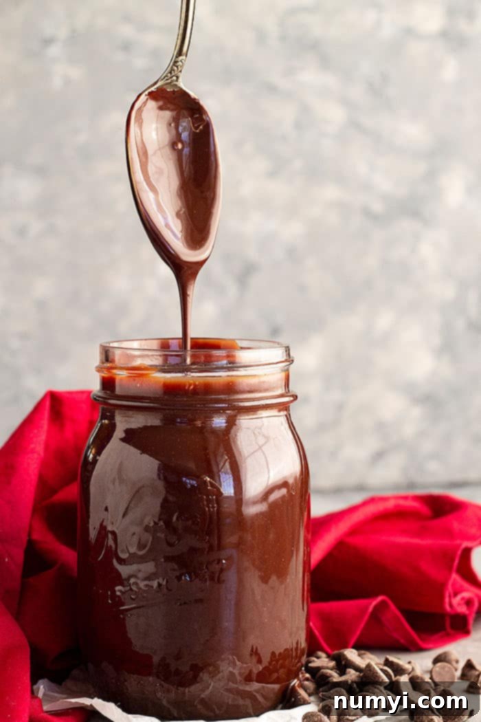 A spoon pulled straight up from a jar of homemade hot fudge sauce letting it drizzle back into the jar, showcasing its smooth consistency.