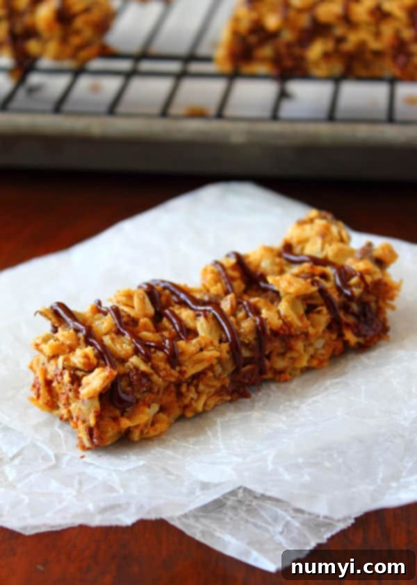 A single Chewy Peanut Butter Granola Bar, drizzled with chocolate, rests on parchment paper, with more bars visible on a cooling rack in the background.