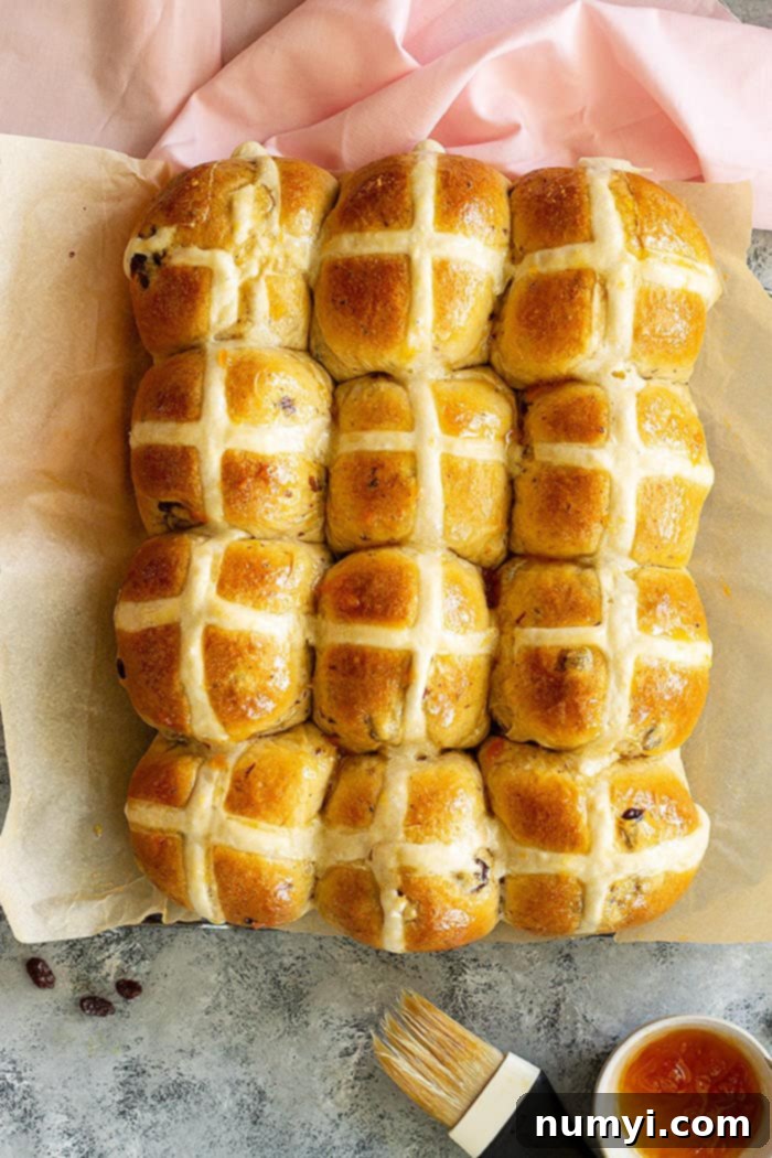 Top down view of a pan of freshly baked hot cross buns, garnished with a white flour paste cross and a shiny apricot glaze.