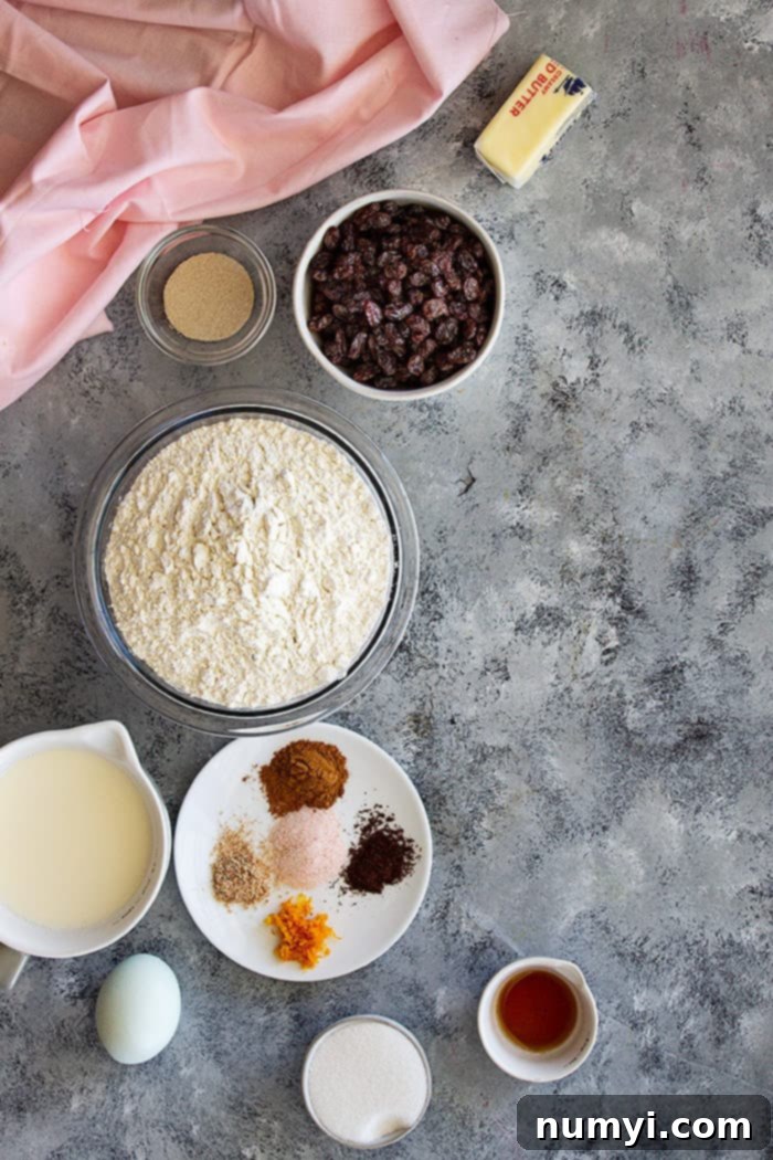 Assortment of fresh ingredients for making hot cross buns, including milk, flour, sugar, eggs, butter, spices, orange zest, and raisins.