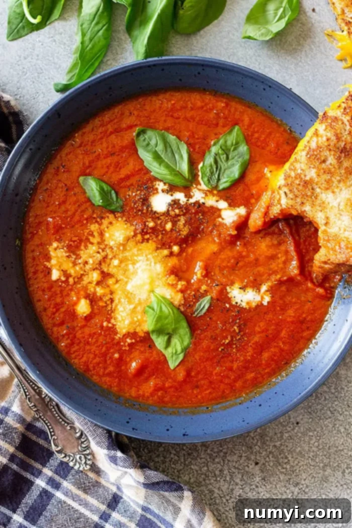 Top down view of a bowl of tomato soup garnished with basil, parmesan cheese, and swirl of cream, and a grilled cheese off to the side. 