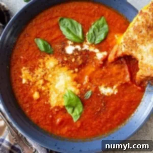 Top down view of a bowl of tomato soup garnished with basil, parmesan cheese, and swirl of cream, and a grilled cheese off to the side.