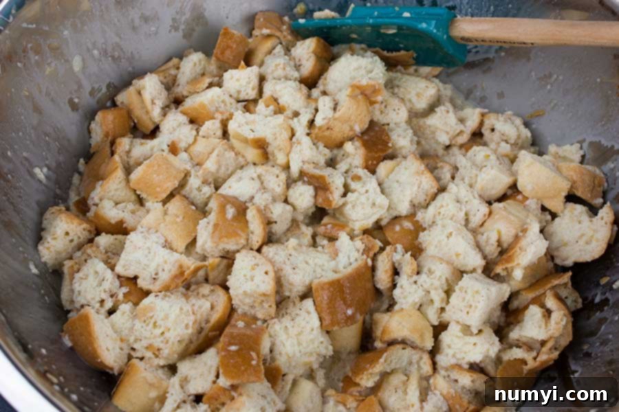 The rich egg mixture being poured evenly over the cubed bread in a large mixing bowl, ensuring every piece is coated.