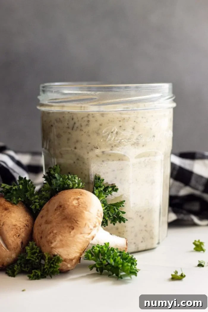 From Scratch Creamy Mushroom Concentrate 2 Homemade condensed cream of mushroom soup in a clear glass jar, with fresh mushrooms and parsley artfully arranged beside it on a wooden surface.
