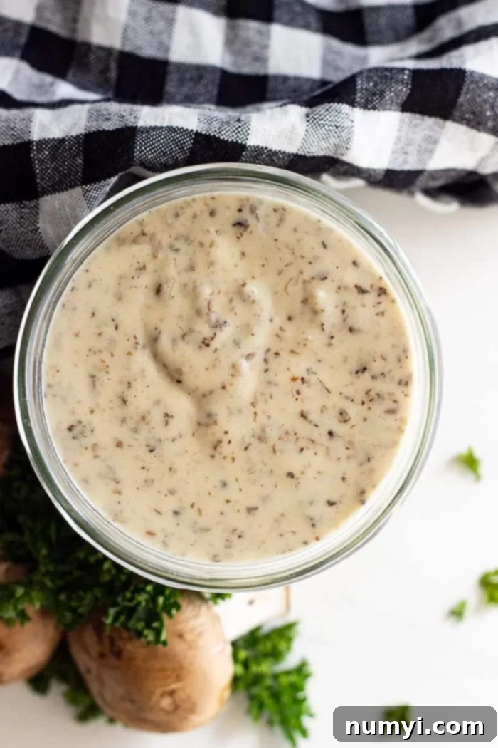 From Scratch Creamy Mushroom Concentrate 6 An inviting overhead view of freshly made homemade condensed cream of mushroom soup, elegantly presented in a glass jar. The jar is surrounded by a charming black and white checkered napkin, fresh mushrooms, and vibrant parsley sprigs, suggesting natural and wholesome ingredients.