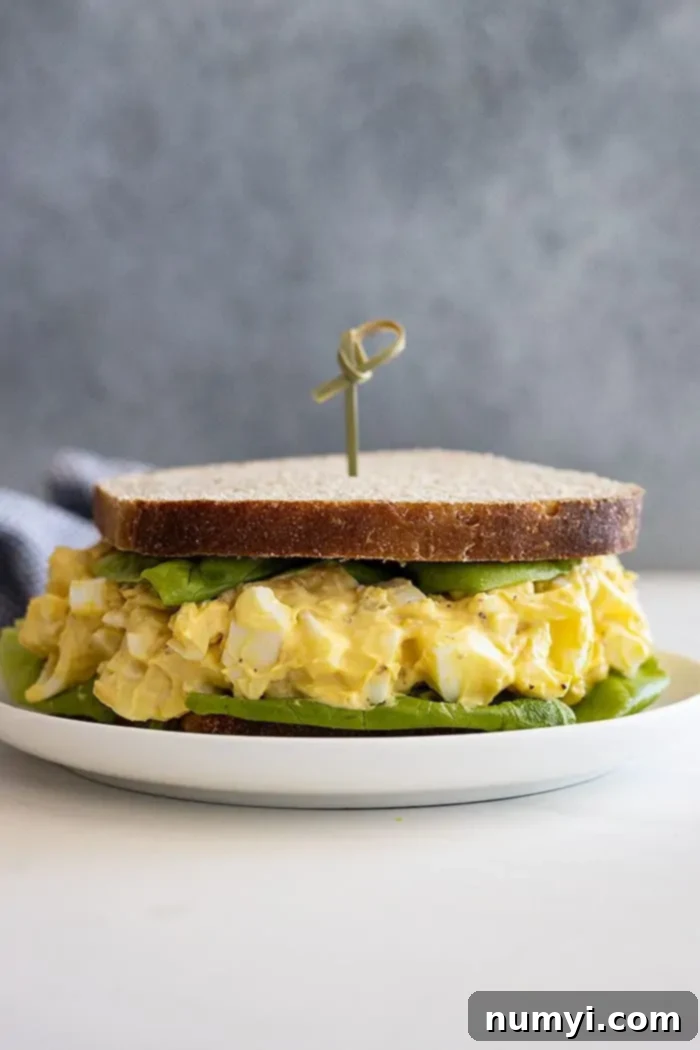 Irresistible Egg Salad 6 A perfectly prepared egg salad sandwich, layered with a large lettuce leaf, presented on a white plate with a decorative toothpick. The focus is on its enticing texture and fresh ingredients.