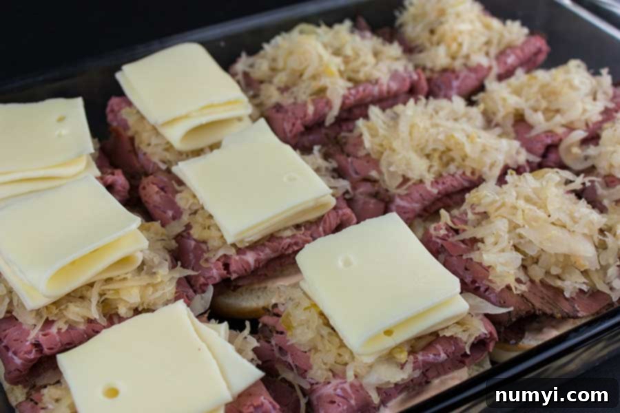 Assembling pastrami and swiss cheese sliders in a baking dish with Russian dressing and pastrami layers.