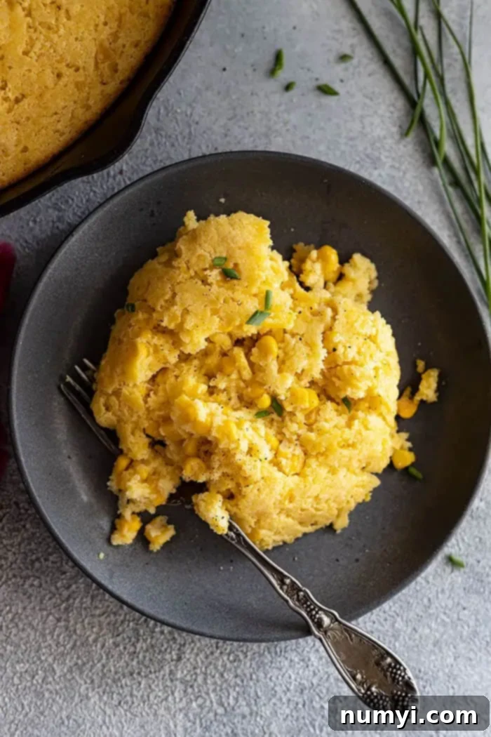 A piece of golden-brown cornbread casserole on a dark gray plate, garnished with fresh chives and a sprinkle of black pepper, viewed from overhead.