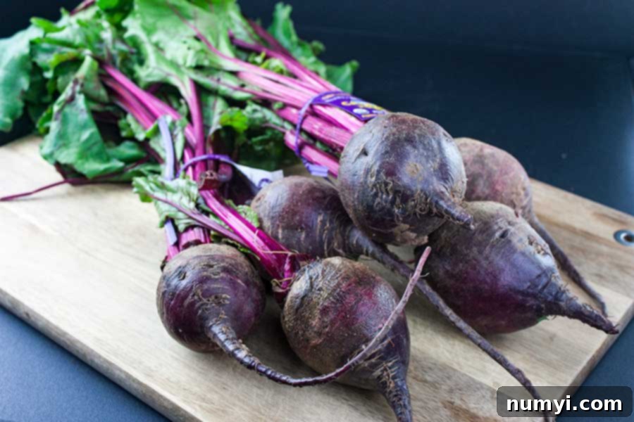Fresh, raw beets with their vibrant green tops still attached, resting on a rustic wooden cutting board, ready for preparation.