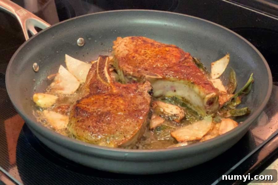 Pork chops cooking in a skillet with a rich butter, onion, garlic, and sage sauce.