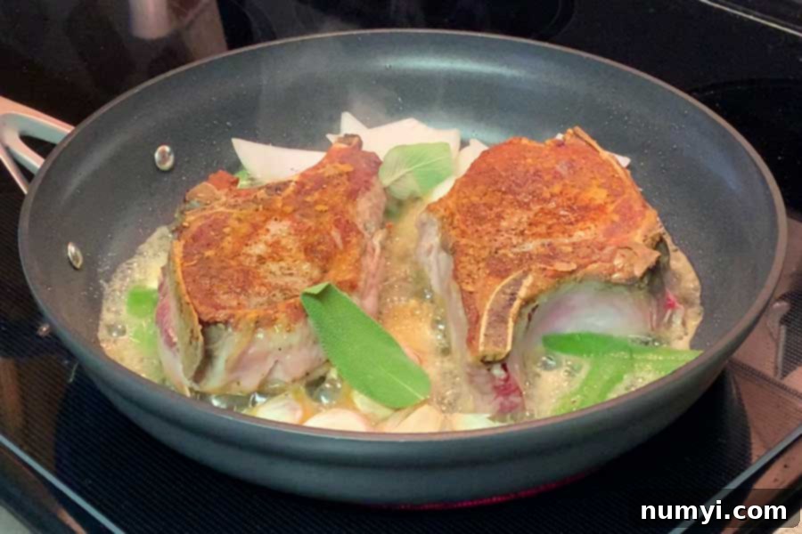 Pork chops cooking in a skillet with fresh sage leaves.