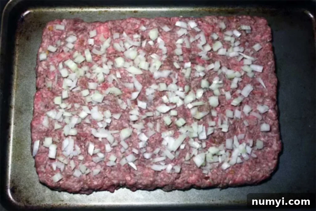 Ultimate Oven Cheeseburger Sliders 3 Raw hamburger meat patted into a thin, even square on a baking tray, generously topped with finely chopped onions, ready for baking.
