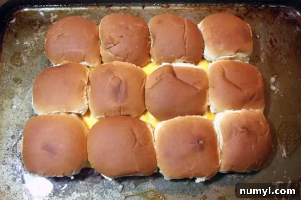Ultimate Oven Cheeseburger Sliders 5 Oven baked sliders on a baking sheet, with the top halves of dinner rolls placed directly on the melted cheese layer, ready for final baking.