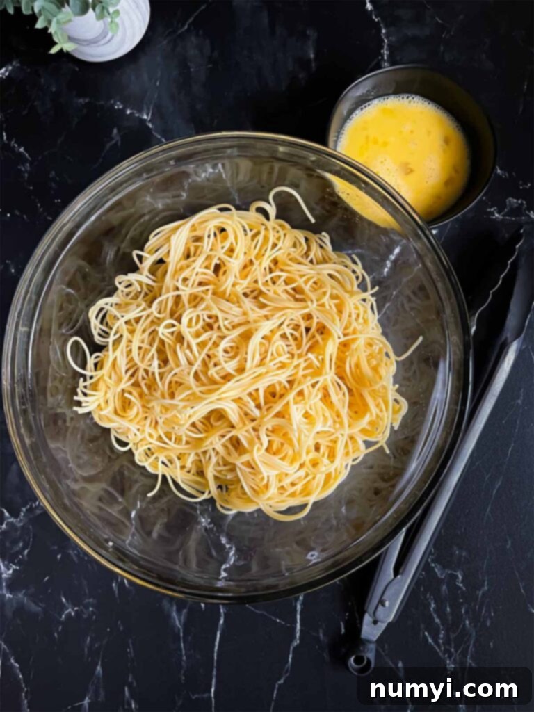 Boiled thin spaghetti in a large glass bowl with the beaten eggs in a small bowl on a dark surface.