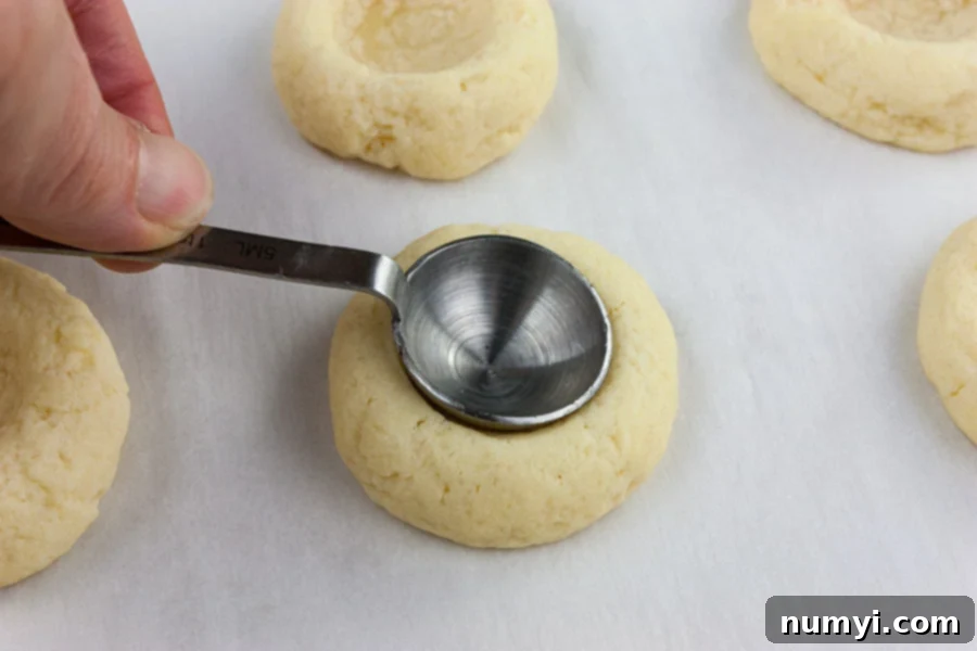 Homemade Jam Thumbprint Cookies 5 A close-up of a baking sheet with partially baked thumbprint cookies, and a teaspoon being used to carefully reform the indentations after their initial bake.