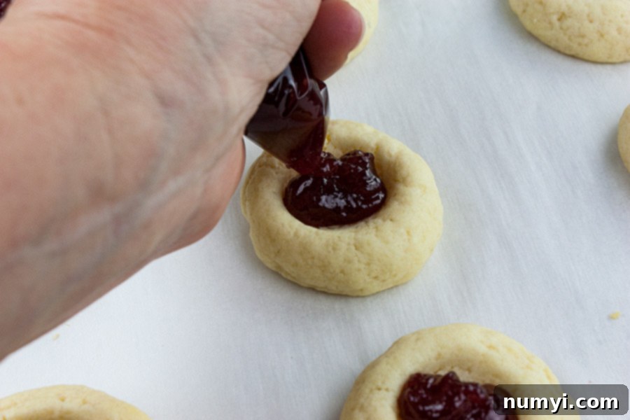Homemade Jam Thumbprint Cookies 6 A hand piping vibrant red raspberry jam into the indentations of partially baked thumbprint cookies on a baking sheet, ready for the final bake.