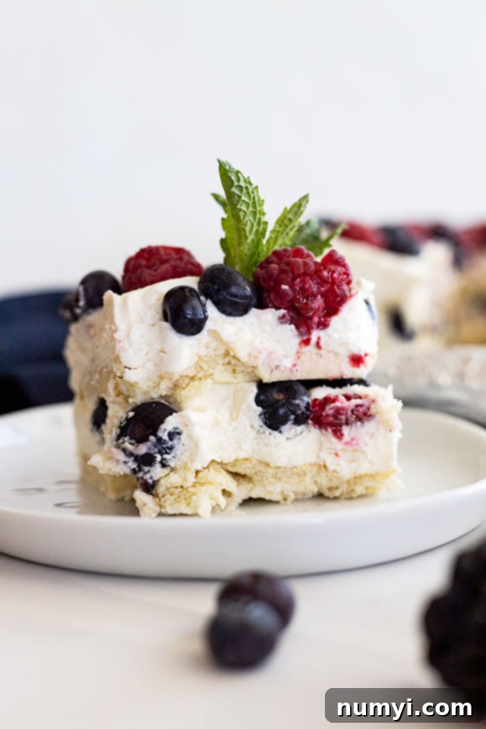 A perfectly sliced piece of berry icebox cake on a pristine white plate, elegantly garnished with a fresh mint sprig and a few extra scattered berries. The distinct layers of creamy filling, softened cookies, and colorful fruit are clearly visible, showcasing the dessert's inviting texture and fresh appeal.