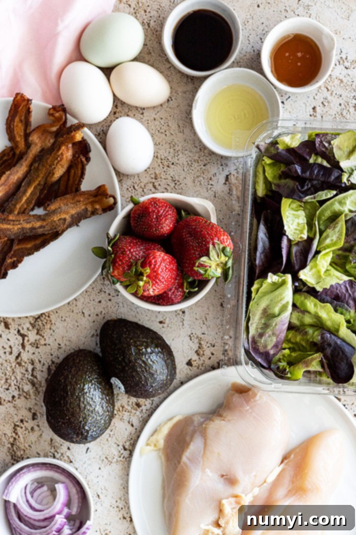 Close-up shot of fresh ingredients laid out for Strawberry Cobb Salad preparation: spring mix, sliced strawberries, grilled chicken, bacon, hard-boiled eggs, avocado, and red onion.