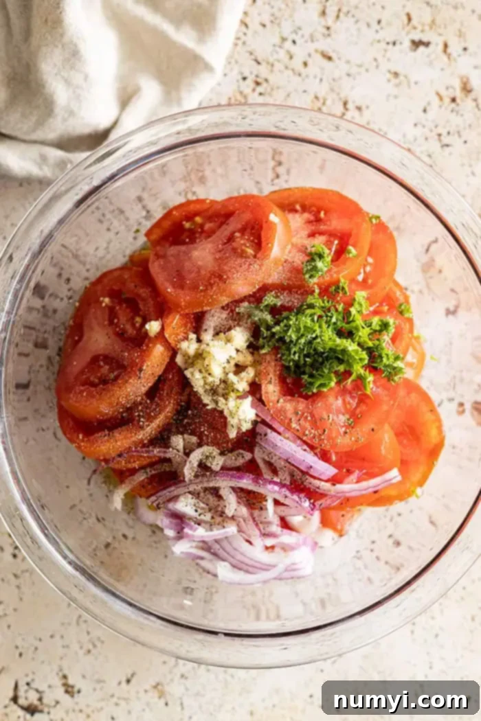 overhead closeup: all the ingredients for this recipe for marinated tomatoes in a glass bowl, ready to be mixed, showing sliced tomatoes, red onion, garlic, and fresh herbs