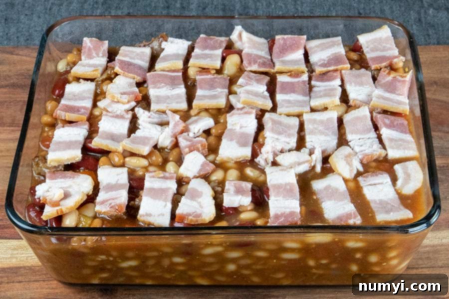 Uncooked baked beans mixture in a glass casserole dish, topped with strips of raw bacon.