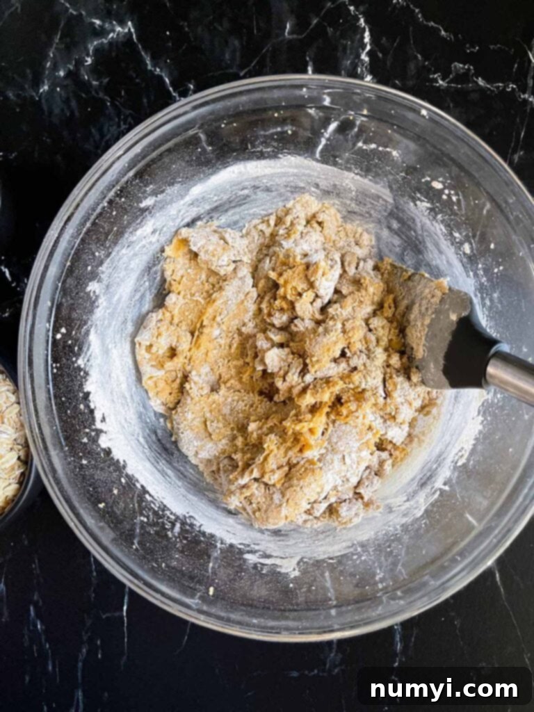 Flour mixture lightly folded into the wet ingredients for oatmeal raisin cookies in a glass bowl, with a few unmixed streaks remaining.